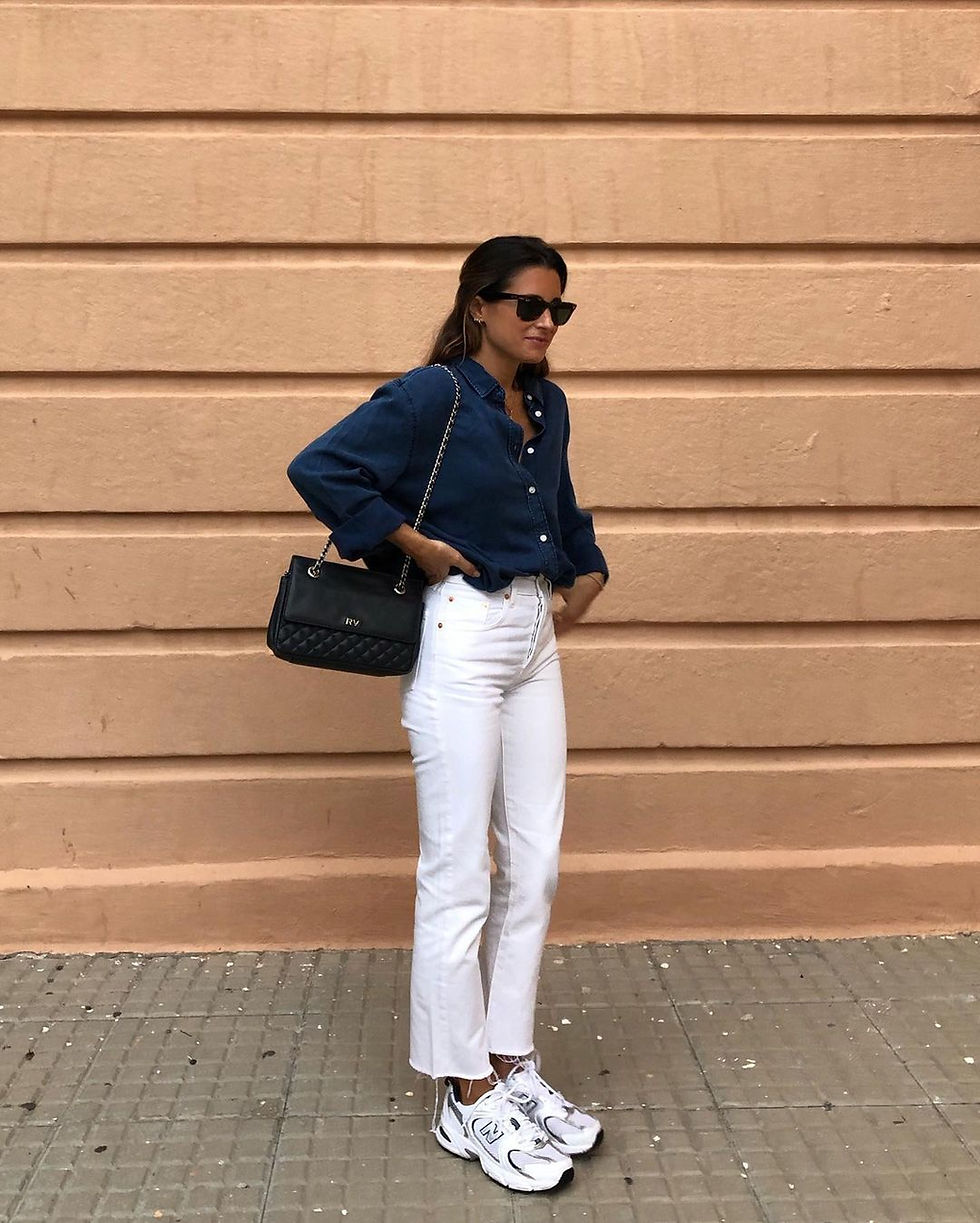 Woman in sunglasses wearing a blue denim shirt, white jeans, and new balance sneakers stands against a peach wall. She holds a black quilted bag.