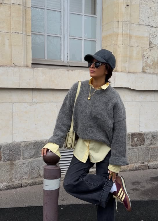 A stylish woman in an urban setting demonstrates how to wear the Spring/Summer 2026 'Retro Sneakers' trend with a chic, layered outfit. She is wearing Adidas-style maroon and yellow gum-sole sneakers with dark denim jeans, a grey knit sweater over a yellow collared shirt, a dark grey baseball cap, sunglasses, and a small cream shoulder bag, showing a wearable way to blend sporty footwear with everyday style.