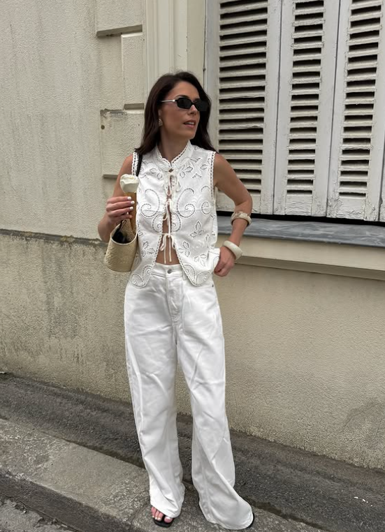 Woman in white embroidered white vest and white jeans, stands by shuttered window, holding ice cream. She wears sunglasses and a woven bag.