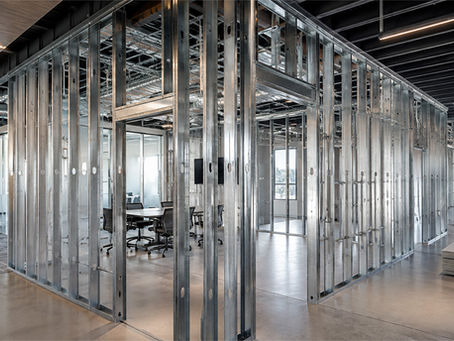 Unfinished modern office interior with exposed metal framing, glass meeting rooms, chairs, and stacked panels under open ceiling.