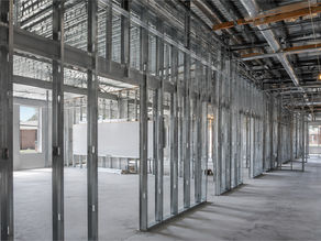 Unfinished school hallway with metal studs, exposed ducts and purple lockers, sunlight through large windows, no people.