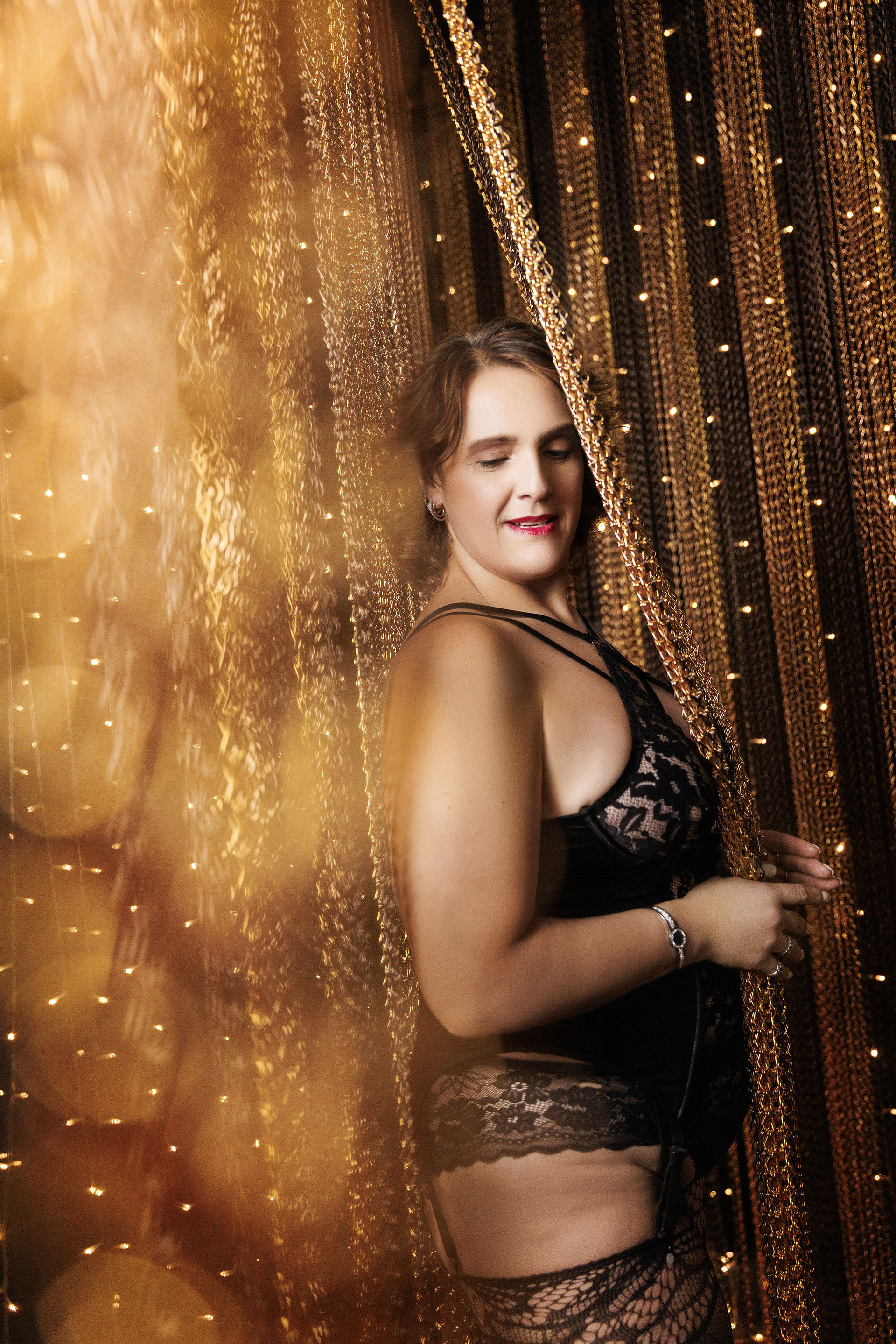 A woman in elegant black lace lingerie stands confidently against a backdrop of shimmering golden chains and warm glowing lights.  She smiles softly, with one hand holding the chains, exuding poise and allure 