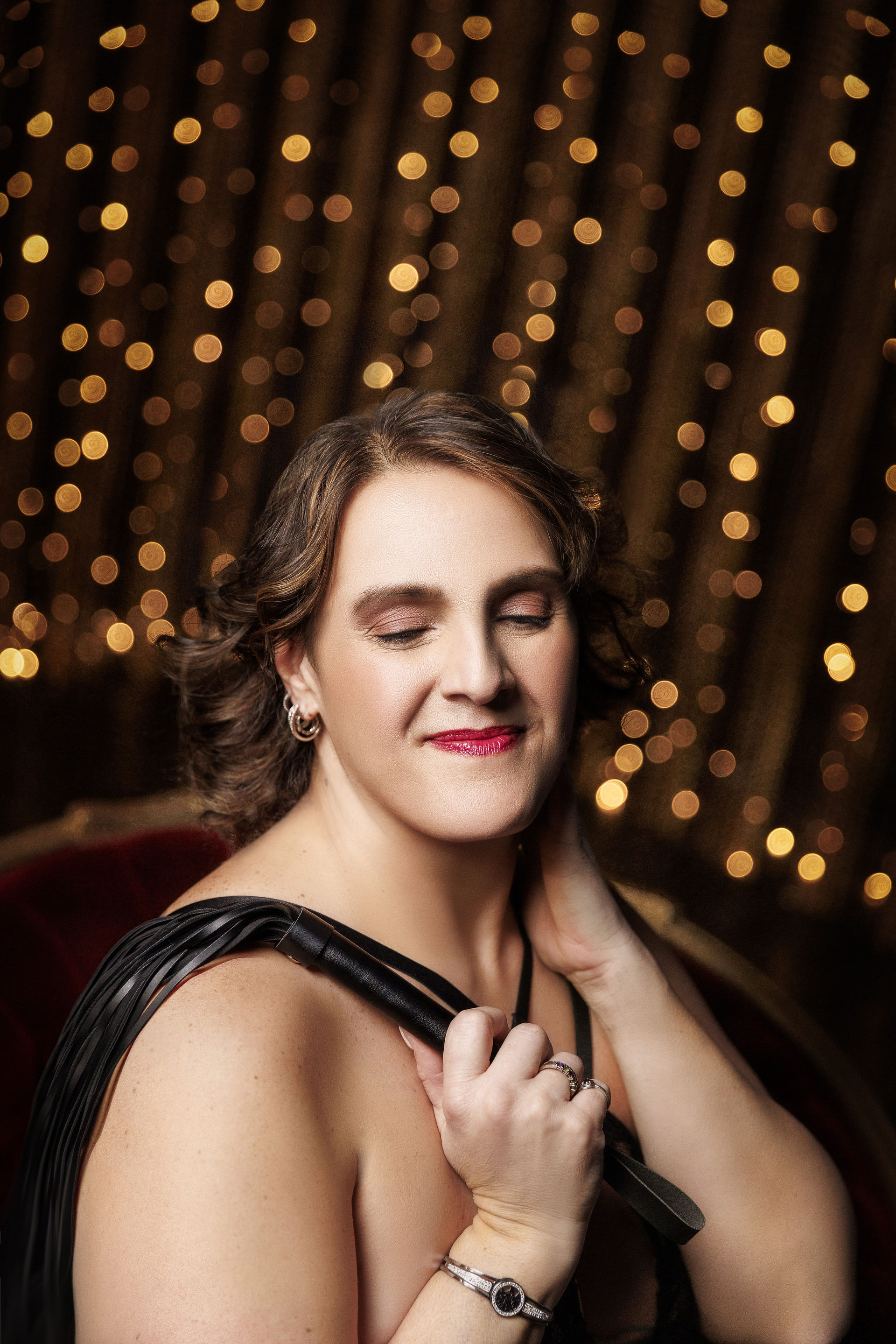 A woman with closed eyes and a serene smile, holding a black leather strap over her shoulder, wearing elegant black lingerie, with golden bokey lights softly glowing in the background, exuding empowerment and self-confidence. 