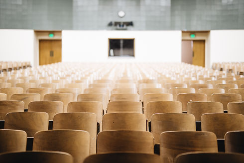 Imagem ilustrativa de uma sala de aula vazia, com inúmeras cadeiras em madeira e uma janela na parte do fundo. A imagem está propositadamente desfocada.