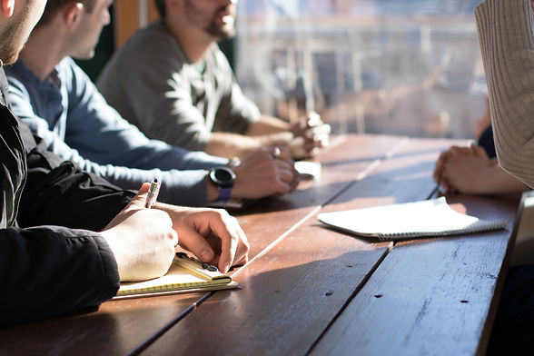 Imagem ilustrativa de pessoas aleatórias tomando notas em cadernos e blocos, sobre uma mesa de madeira iluminada por raios de sol. Há 3 pessoas visíveis parcialmente na imagem, das quais 2 são homens brancos.