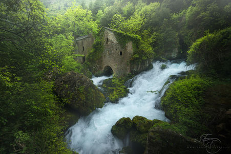 Waterfall rushing past mossy rocks beside old stone ruins in a green forest