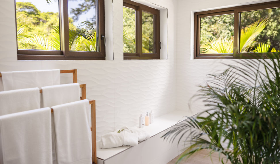 Modern resort bathroom with textured white walls, a wooden towel ladder, and windows looking out at tropical plants at The Hacienda Resort