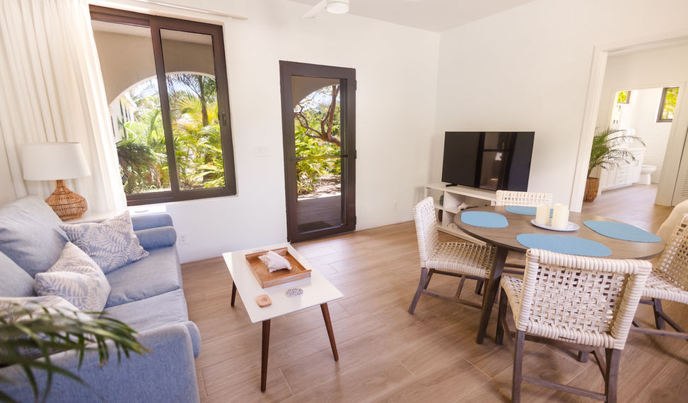 Modern resort living room with a light blue sofa, coffee table, flat screen TV, and glass doors opening to tropical gardens at The Hacienda Resort
