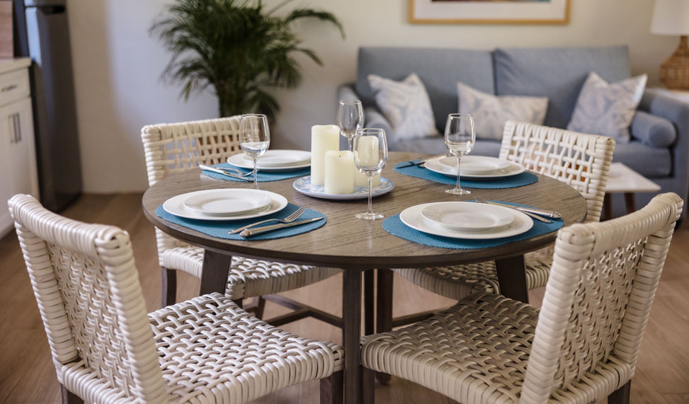 A round wooden dining table set for four with white plates, wine glasses, and woven chairs in a luxury resort apartment living space at The Hacienda Resort