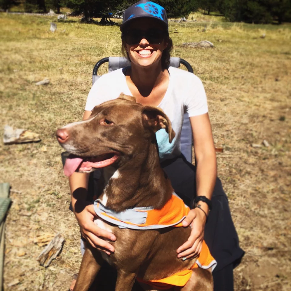 woman smiles while sitting in chair and holding a brown dog wearing an orange vest