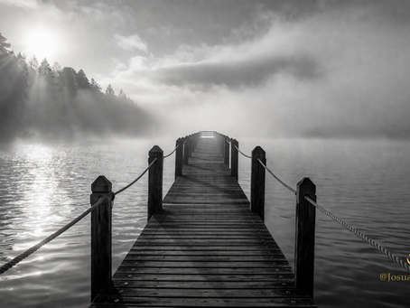 Schwarz-weißes Bild eines langen Holzstegs, der in einen nebelverhangenen See führt. Sanfte Lichtstrahlen durchbrechen den Nebel und erzeugen eine ruhige, meditative Atmosphäre, die Stille, Bescheidenheit und innere Einkehr symbolisiert.