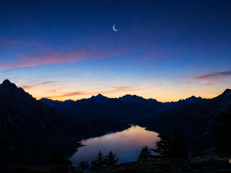 Schmale Neumondsichel über einem stillen Bergsee in der blauen Dämmerung, umgeben von dunklen Bergsilhouetten, zarten rosafarbenen Wolken und der goldenen JosuasGarden-Signatur.