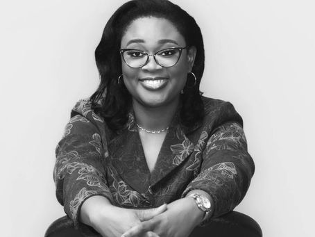 Smiling woman in glasses and floral-patterned jacket leans on chair back. Black and white image with a neutral background.