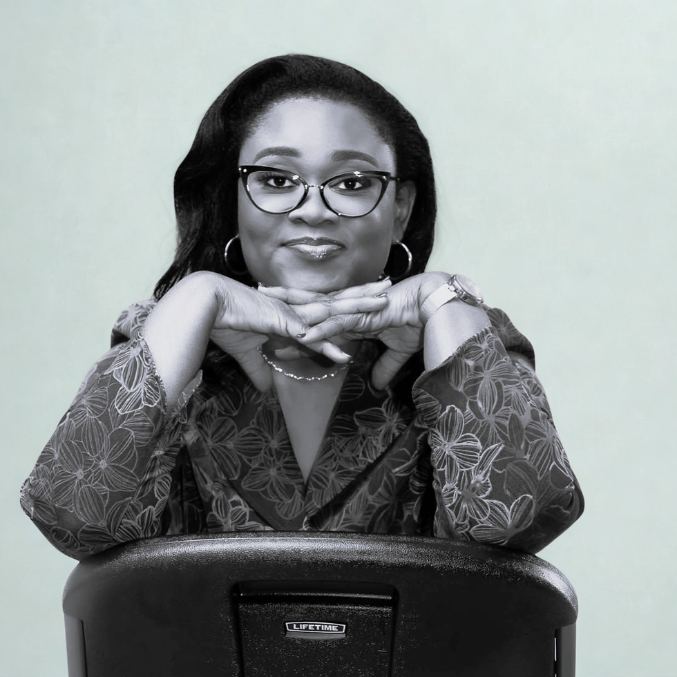 Woman with glasses and patterned blouse leans chin on hands, smiling. Background is light gray; chair has "LIFETIME" text. Relaxed mood.