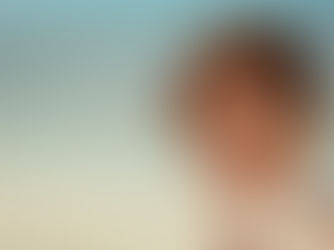 Young woman in her 20s standing by the beach with the ocean behind her, smiling softly into the wind.