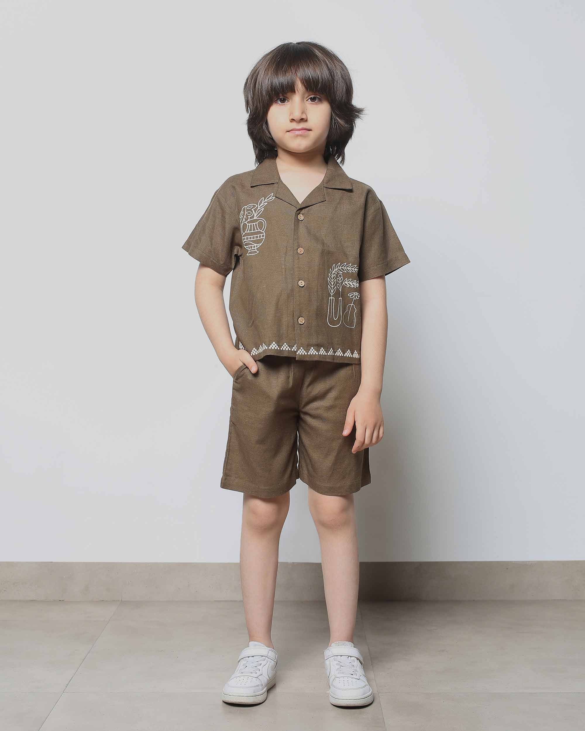 A young boy models a brown two-piece co-ord set with a short-sleeved button-down shirt and shorts featuring a subtle print.