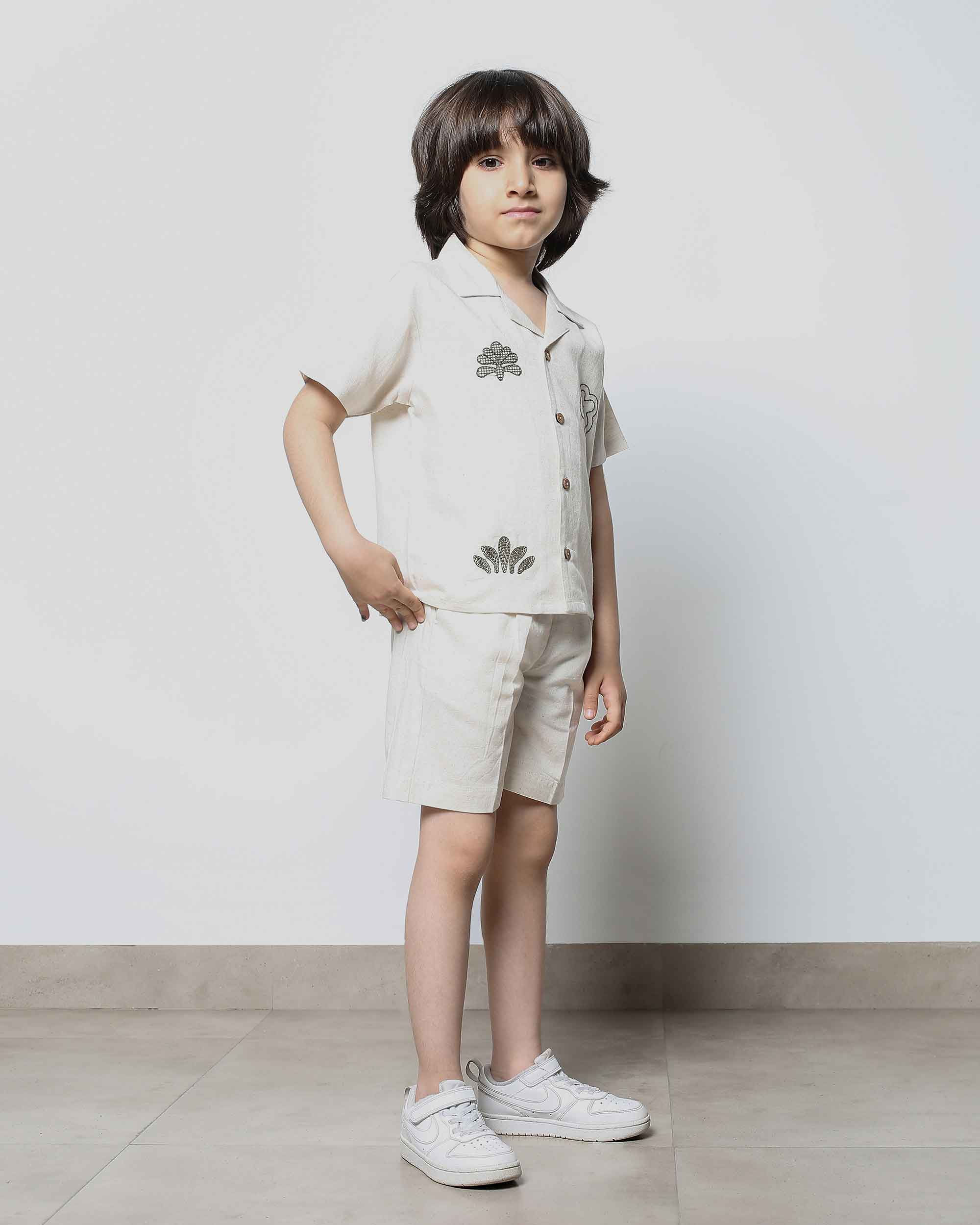 A young boy models a light beige two-piece co-ord set with a short-sleeved button-down shirt and shorts featuring floral embr