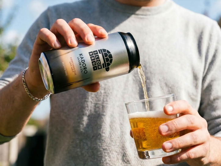 Slow Bru Kadoro craft lager in a can which is being poured into a glass on a sunny day by a mid 30's man in a grey TShirt