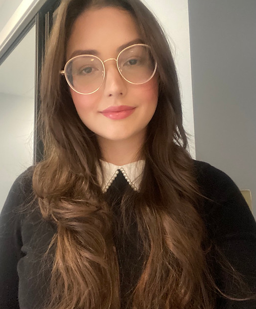 imagem de mulher branca com cabelos lisos castanhos divididos ao maio sobre blusa preta. Usa oculos redondos de grau, dourado