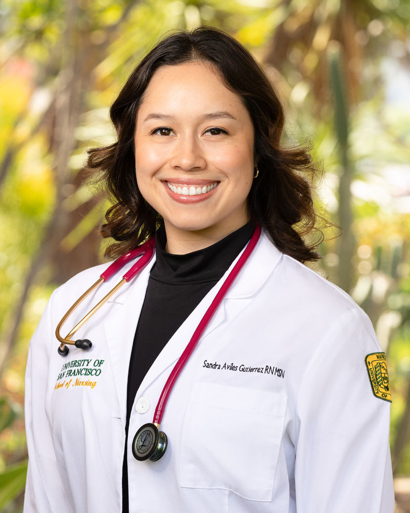 Professional outdoor healthcare headshot of a smiling woman with shoulder-length dark brown hair, wearing a white medical coat over a black top with a stethoscope around her neck. Photographed by Galvez Photography against a softly blurred, tree-lined background, highlighting professional healthcare attire and a confident, approachable expression.
