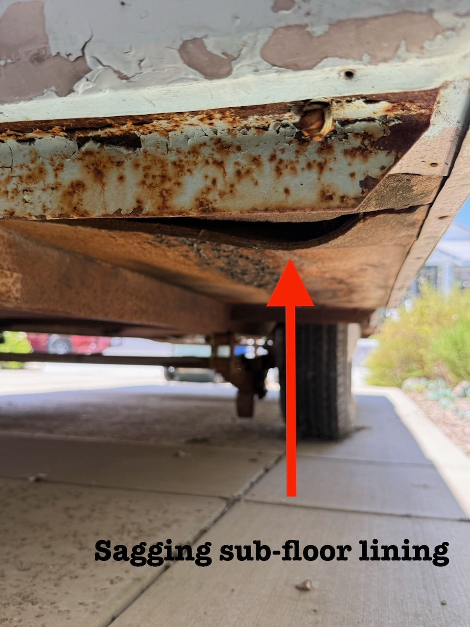 Sagging sub-floor lining under a rusted structure. A red arrow points at the damage. Text reads "Sagging sub-floor lining." Outdoors setting.