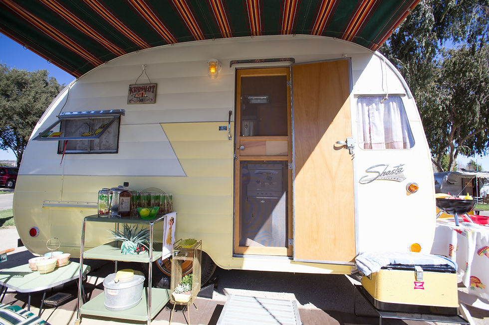 Retro Shasta trailer with striped awning, outdoor setup with drinks and plants, and a sign saying "KampALot." Bright, sunny day.