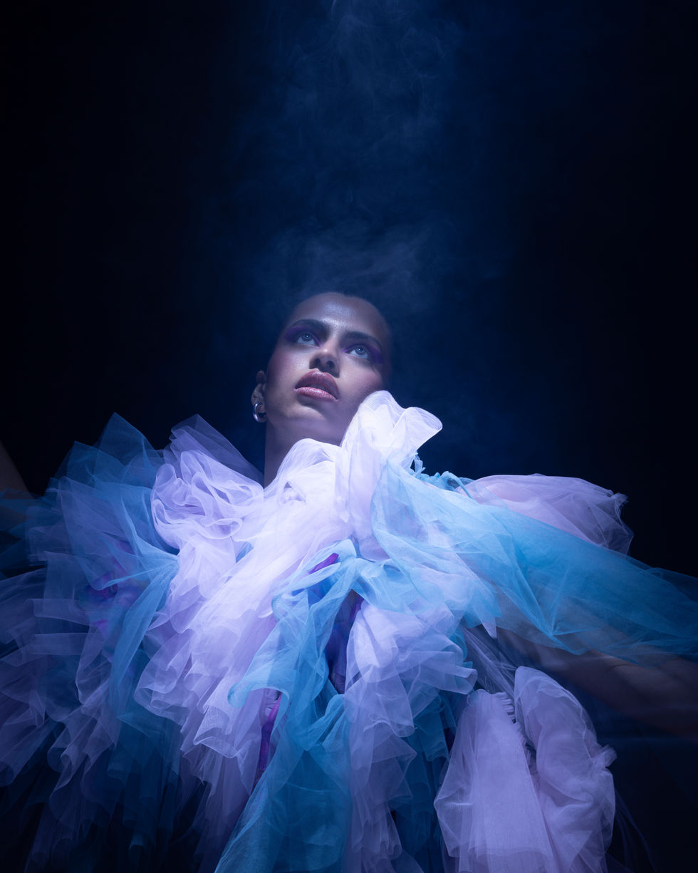 Fine art fashion portrait of a person draped in layers of pastel tulle with eyes closed, surrounded by soft blue smoke, creating an ethereal and dreamlike atmosphere.