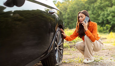 Worried young European woman talking on cellphone and checking her car flat tire on the si
