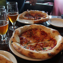 Two pizzas sitting on a black table with two glasses of half-full beer on the left