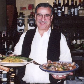 A smiling man wearing a white shirt, black vest, with each hand holding a bowl of food