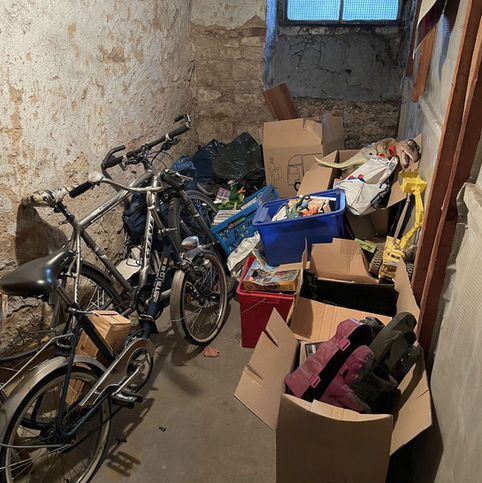 Ein vollgestellter Keller im Altbau.
Schmutz, Staub, alte Sachen, Fahrräder, Müll und Kartons häufen sich. Im Hintergrund ein kleines, milchiges Kellerfenster.