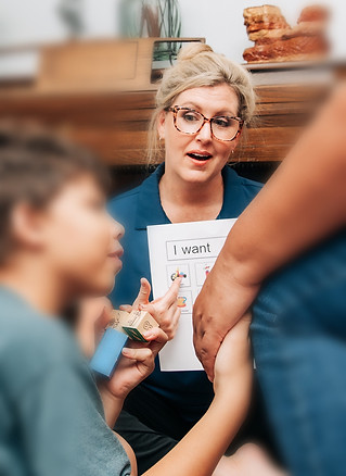 Woman demonstrating a picture communication board to a young buy who is playing blocks and holding his mom's hand.
