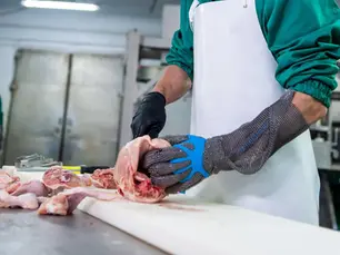 Worker in green attire and white apron cuts raw meat on a table, wearing a chainmail glove. Industrial kitchen setting in the background.