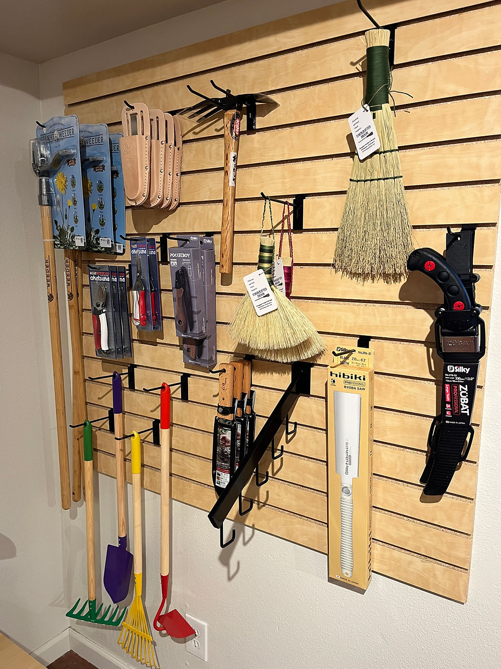 Wall-mounted display of gardening tools, including rakes, saws, and brooms. Colorful handles and packaging, wood panel background.