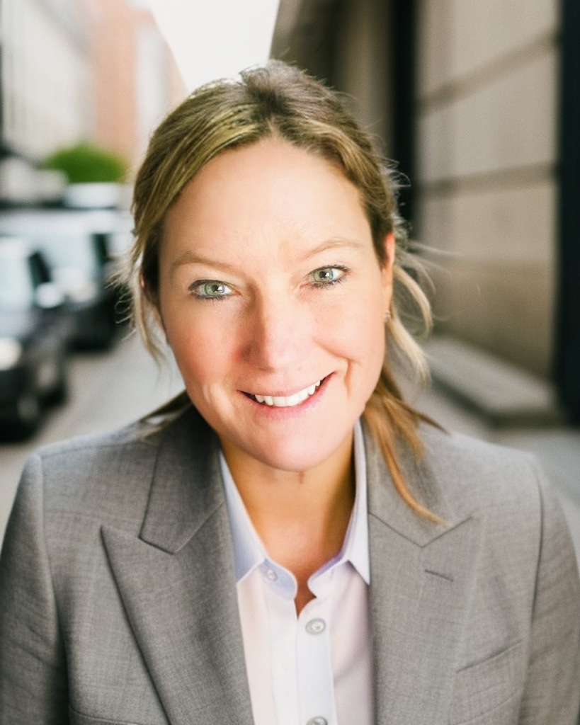 Smiling woman in a gray suit on a city street. Urban background with blurred buildings and vehicles. Bright, welcoming mood.