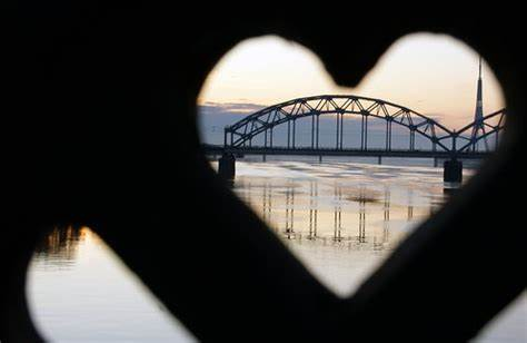 Heart-shaped frame view of a steel bridge at sunset, reflected on calm water. Sky has soft pastel hues, creating a serene mood.