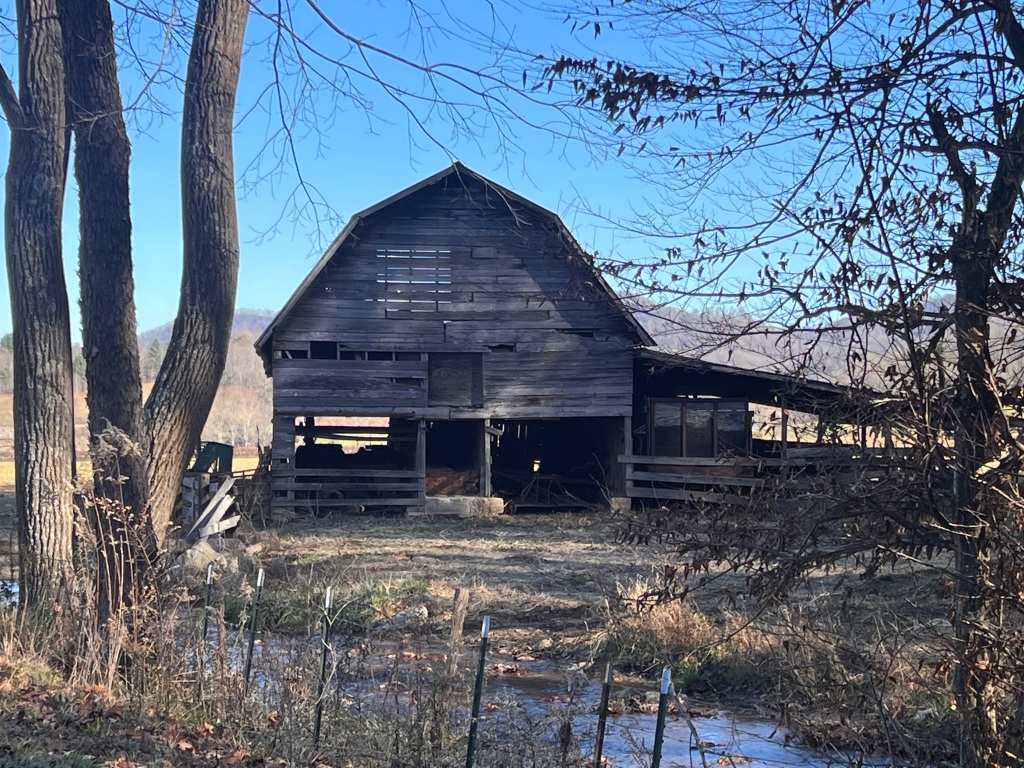 Old wooden barn in a rural field with trees and a clear blue sky. Weathered wood, barren branches, and a small creek create a serene setting.