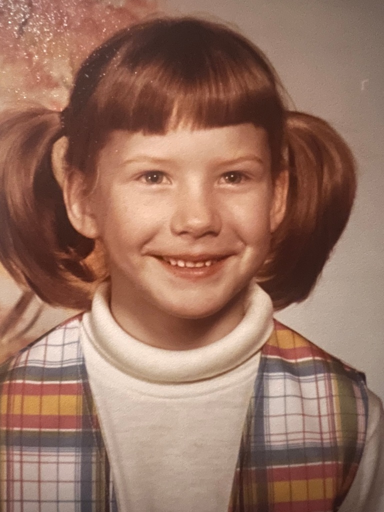 Young girl with pigtails and a smile, wearing a colorful plaid vest over a white turtleneck, poses in front of a soft, blurred background.