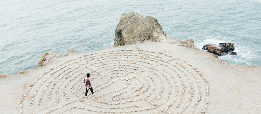 A woman walking slowly through a stone labyrinth path, surrounded by nature, pausing with intention and reflection.