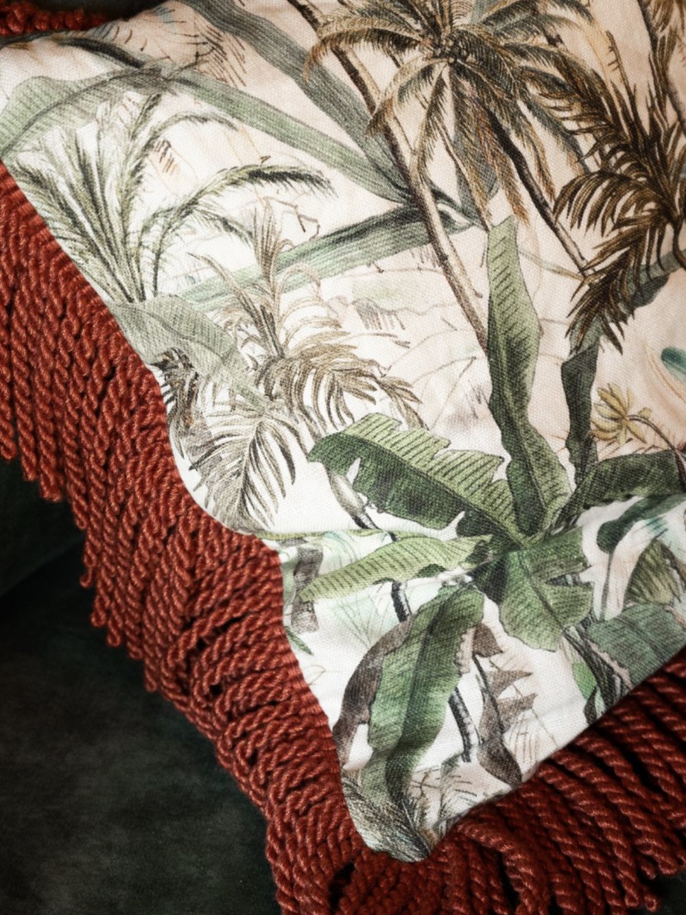 Coussin en tissu palmiers bordé de franges terre cuite sur une banquette en velours vert