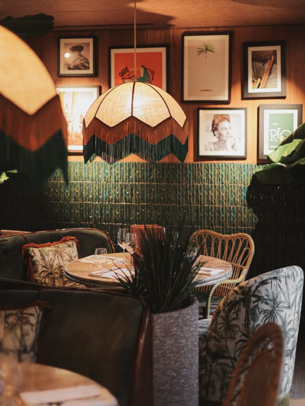 Le mur de cadres du Poncho, suspension à franges, banquette en velours vert et fauteuil en tissu palmiers