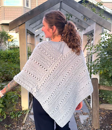 Person wearing a light blue crocheted wrap with textured stitches, shown from the back while standing on a garden walkway