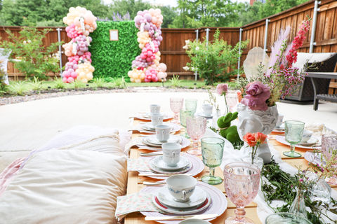 A tea party on picnic tables facing a greenery wall backdrop with balloons