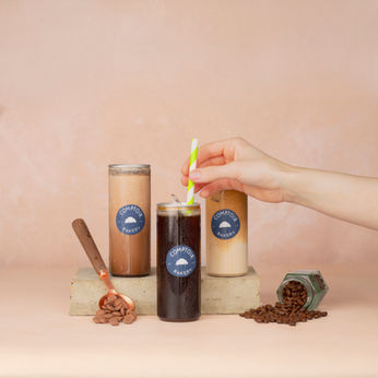Three iced coffee drinks, straw, coffee beans, logo on label, hand holding straw, beige background.