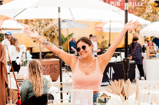 Woman with arms raised smiles at Dreamers Markets, selling jewelry at booth.