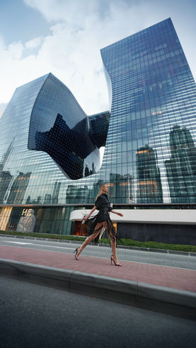 A woman in a stylish black outfit confidently strides along a sidewalk in front of a modern skyscraper with a unique, curved design. The tall glass building reflects the city skyline, creating a dynamic urban backdrop.