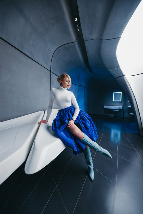 A woman in a sleek, modern interior sits on a white bench, wearing a white long-sleeve top, a vibrant blue skirt, and matching blue boots, with her hair styled in an updo.