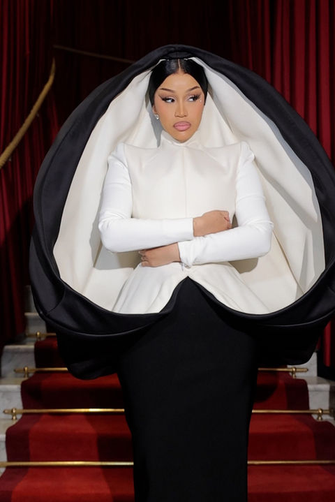 A woman in an elegant white satin dress poses gracefully against a richly decorated wooden door, surrounded by marble columns. She wears sparkling jewelry and strappy high heels, exuding a sophisticated style.