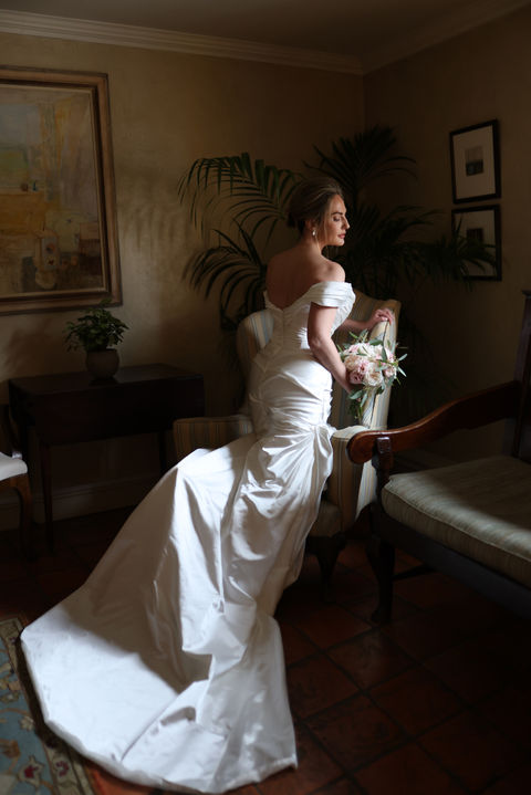 A bride in an elegant white gown sits on a chair, holding a bouquet of pink and white flowers. The room is warmly lit, with a painting on the wall and lush greenery in the background.