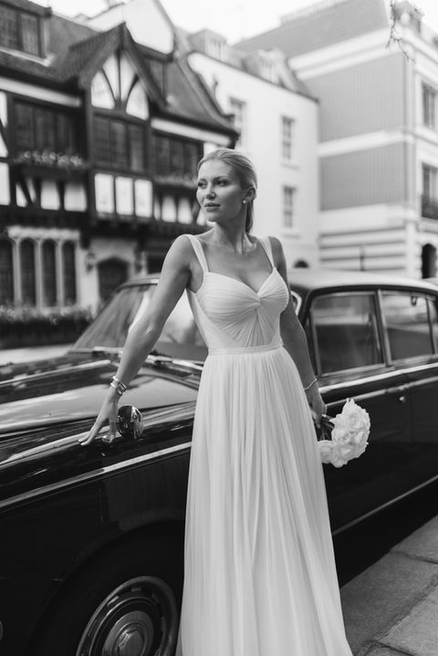 A woman in a flowing white dress stands elegantly beside a classic black car, holding a bouquet of flowers, with charming, vintage buildings in the background.